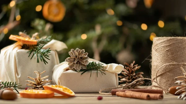 Концепция нулевого расточительства Рождества. Упакованные в натуральные ткани подарки и украшения из натуральных материалов на деревянном столе возле елки с огнями — стоковое фото