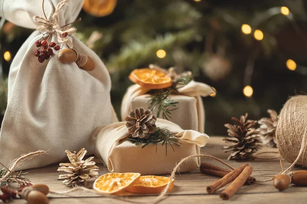 Концепция нулевого расточительства Рождества. Упакованные в натуральные ткани подарки и украшения из натуральных материалов на деревянном столе возле елки с огнями — стоковое фото