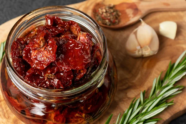 Sun-dried tomatoes with garlic, rosemary and spices in a glass jar on an olive wood cutting board — Stock Photo, Image