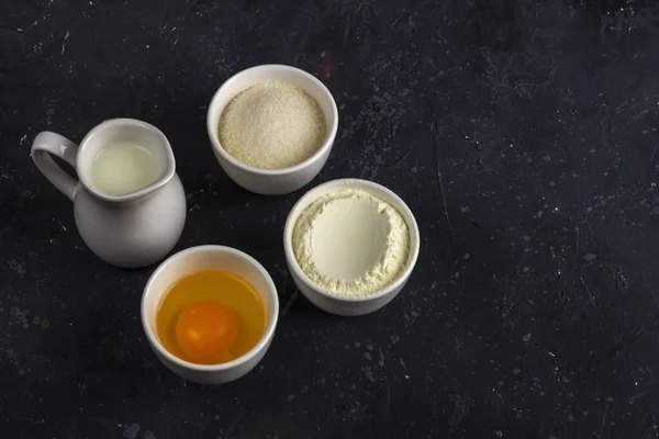 Baking background. Ingredients for cooking cake (flour, egg, sugar, milk) in bowls on dark table. Food concept. Close up layout, copy space for text.