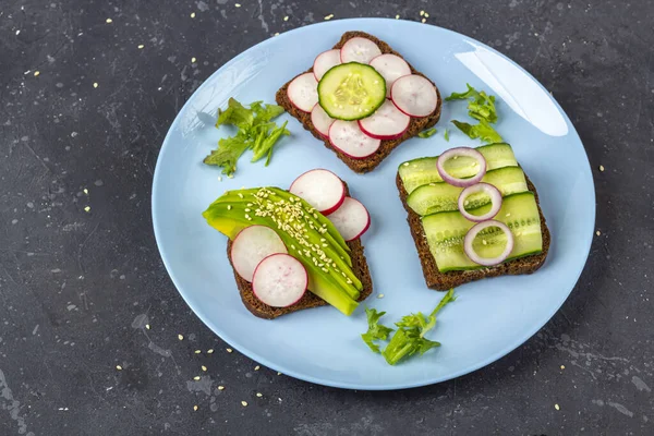 Superfood open vegetarian sandwich with different toppings: avocado, cucumber, radish on plate on dark background. Healthy eating. Organic and veggie food. Close up, copy space for text