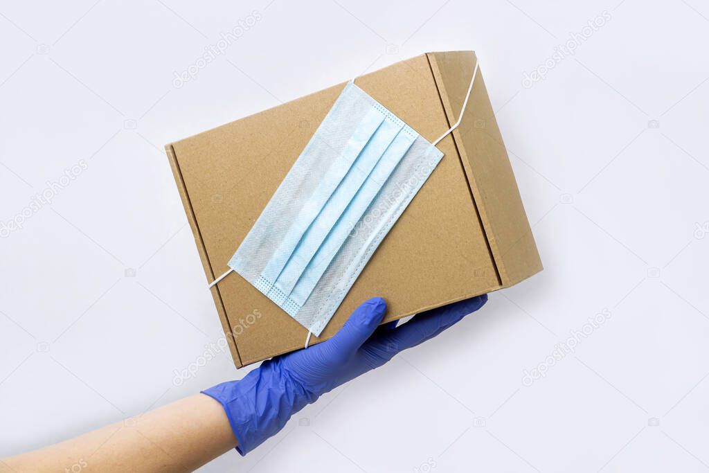 Safe contactless Delivery service during quarantine. Young woman hand in rubber holds box with protective medical mask. Stay at home, online shopping during coronsavirus outbreak. Copy space, mock up