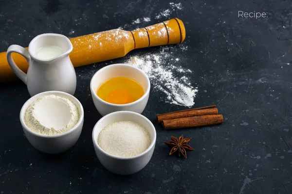 Baking background. Ingredients and utensils for cooking cake (flour, egg, milk, sugar, rolling pin, wooden spoon) on dark table. Food concept. Close up layout, copy space for text.