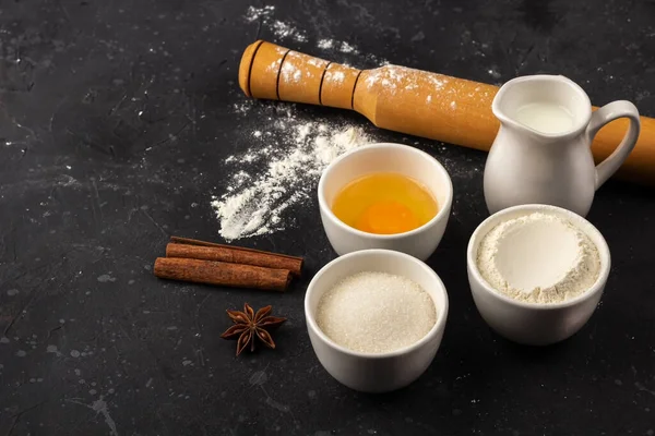 Baking background. Ingredients and utensils for cooking cake (flour, egg, milk, sugar, rolling pin, wooden spoon) on dark table. Food concept. Close up layout, copy space for text.