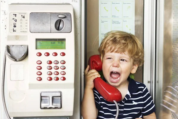 Ребёнок разговаривает по стационарному телефону. Мальчик в телефонной будке. Красный телефон — стоковое фото
