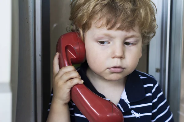 Baby talking on landline phone. A boy in a telephone booth. Red handset