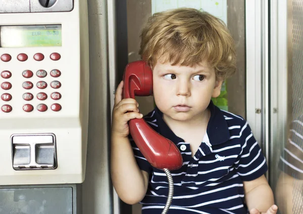 Baby talking on landline phone. A boy in a telephone booth. Red handset