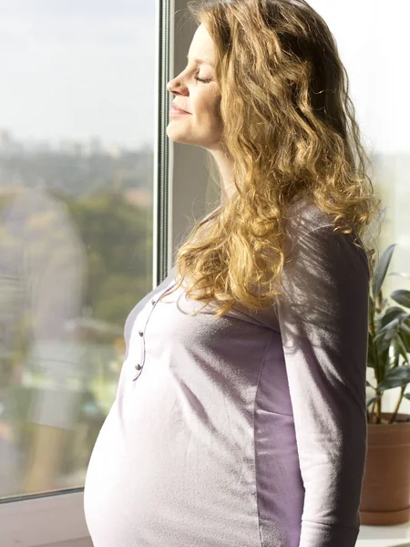 Schöne schwangere Blondine steht am Fenster und genießt die Sonne — Stockfoto