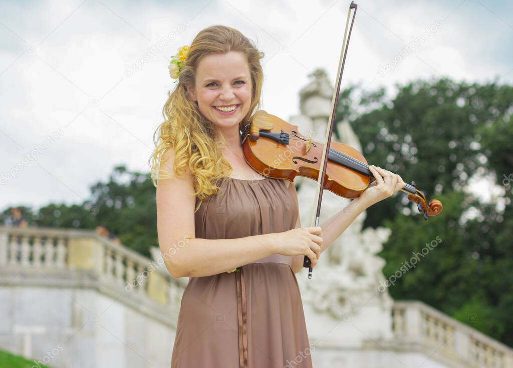 Beautiful smiling girl playing the violin outdoors. Curly blonde in a long dress. Girl playing the violin