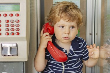 Ciddi bir çocuk telefonda konuşuyor. Telefon kulübesindeki çocuk. Elinde kırmızı bir cep telefonu olan küçük bir çocuk.