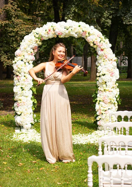 Bride in the park. Young woman with a violin on the background of the wedding arch. Beautiful girl with a violin outdoors.