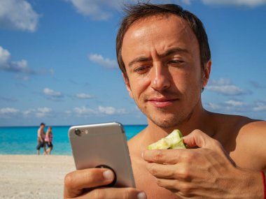 Bir adam armut yer ve sahilde akıllı telefon kullanır. Bilgisayar bağımlılığı.