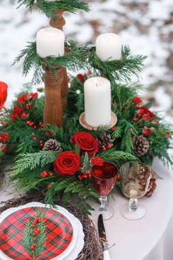  Çam dalları, ladin kozalakları, kırmızı güller, turuncu kızılcık böğürtlenleri ve ahşap şamdanlarda beyaz dekoratif mumlar. Şarap bardakları. Beyaz bir tabakta İskoç kareli bir süs ve dallardan bir süs var.. 