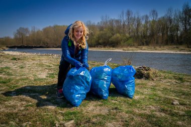 Sarı saçlı, mavi eldivenli genç bir kadın güneşli bir baharın ilk günlerinde plastikten büyük mavi çöp torbalarına kadar nehri temizliyor.