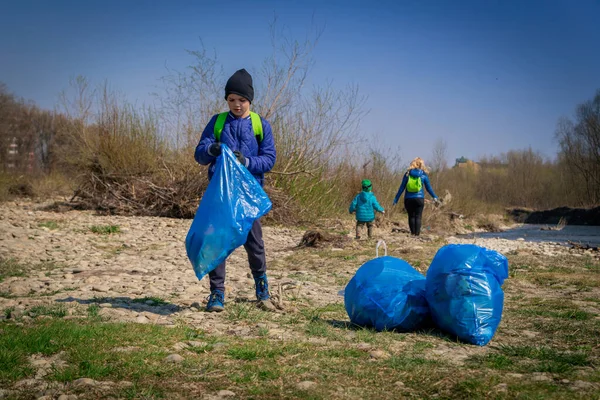 Oğulları mavi eldivenli genç sarışın kadın güneşli bir baharın ilk günlerinde büyük mavi çöp torbalarıyla nehir kıyısını plastikten ve çöpten temizliyor.