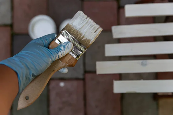 paint brush covered with white paint in hand and paint can paint wooden planks on the dark background in the yard
