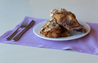 Beyaz tabakta çikolata ve bademli kruvasanlar ve mor peçeteler.