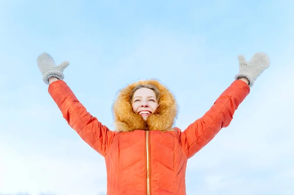 Außenporträt eines lächelnden Mädchens in winterlicher Daunenjacke, hebt die Hände gegen den blauen Himmel — Stockfoto