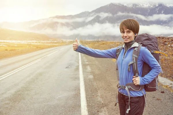 Mujer viajera haciendo autostop. Hermosa joven autoestopista por la carretera durante el viaje de vacaciones en las montañas al atardecer — Foto de Stock