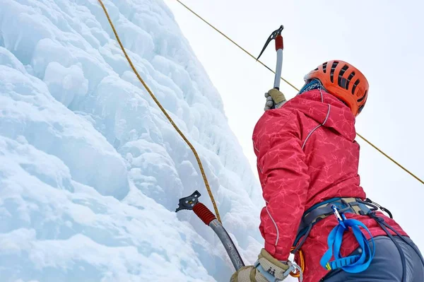 Femme alpiniste avec piolet à outils en casque orange escaladant un grand mur de glace. Portrait de sport extérieur — Photo