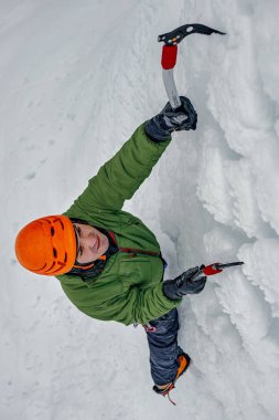 Elinde buz aletleri olan dağcı adam turuncu kasklı baltayla büyük bir buz duvarına tırmanıyor. Açık hava Spor Portresi.