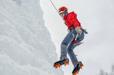 Alpinist woman with ice tools axe in orange helmet climbing a large wall of ice. Outdoor Sports Portrait. clipart