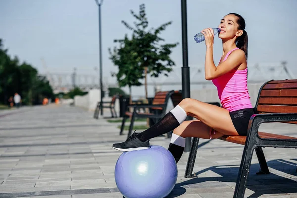 Mulher fitness com garrafa e bola de borracha. Água potável feminina após treinamento em banco no parque de verão — Fotografia de Stock