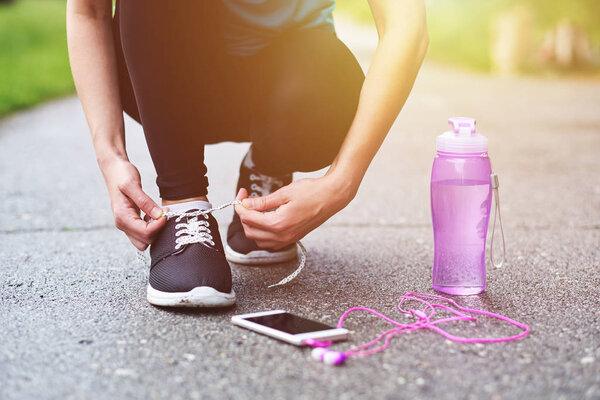 runner woman tying running shoes laces getting ready for race on asphalt with bottle of water and phone with headphones