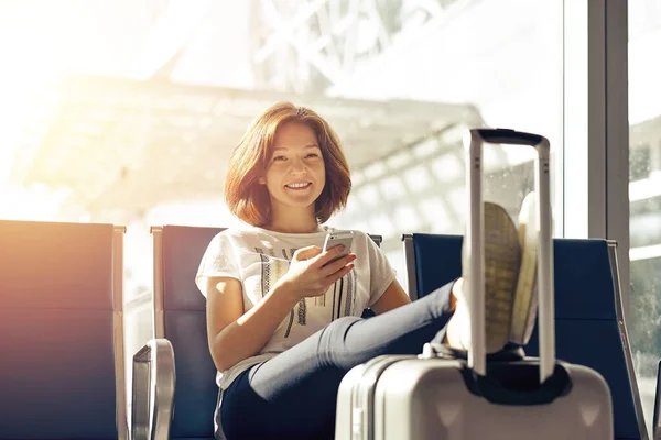 Smiling woman with ticket and baggage in airport. Air travel concept with young casual girl sitting with hand luggage suitcase at gate waiting in terminal