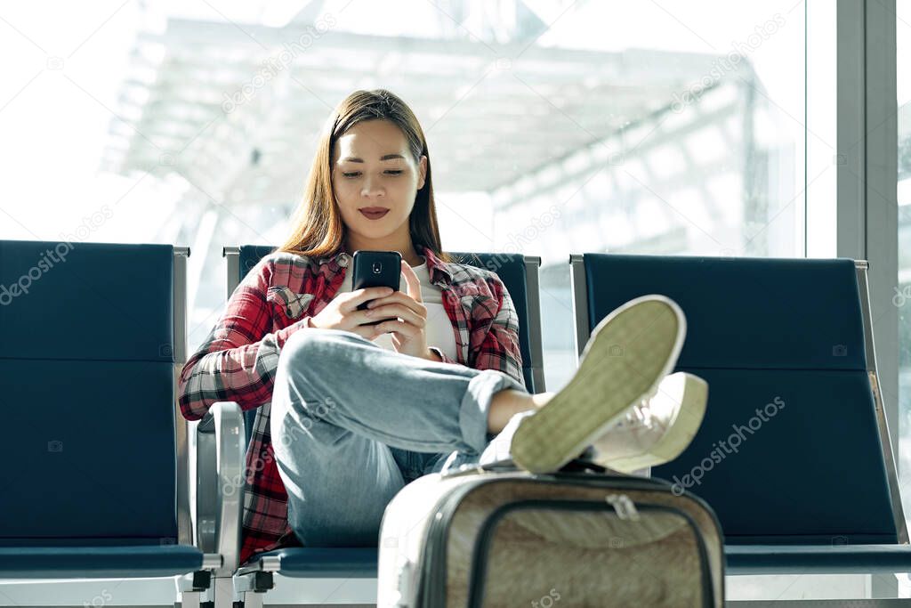 Air travel concept with young casual girl sitting with hand luggage suitcase. Airport asian woman on smart phone at gate waiting in terminal.