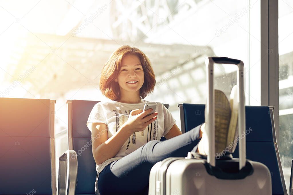 Smiling woman with ticket and baggage in airport. Air travel concept with young casual girl sitting with hand luggage suitcase at gate waiting in terminal