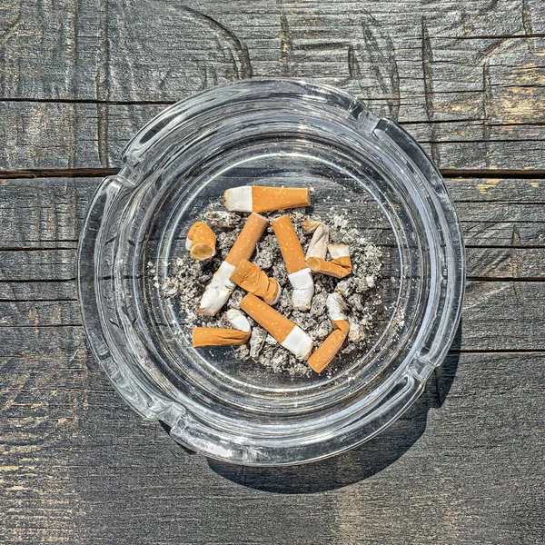 Glass ashtray with cigarette butts and ash on a wooden board. Glass bowl and receptacle with stubbed out cigarettes and ash, outdoor and in the sunlight on a weathered gray wooden table plate. Photo.