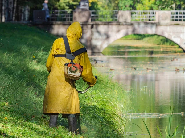 A man in a yellow raincoat mowing the grass next to the pond. Lawn mower at work. A man with a lawn mower. Mowing the grass trimmer. Mowing the lawn.