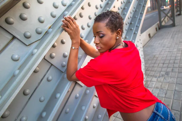 Woman in a red blouse is posing next to bridge. Dark-skinned girl leaned on the base of the bridge. Woman posing on a background of city streets. African in a red T-shirt. Girl with a short haircut