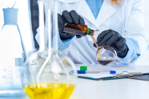Lab technician pours dark liquid into a flask. Concept - a laboratory career. Concept - production of prescription drugs. The doctor in protective gloves mixes the ingredients. The study. Analysis