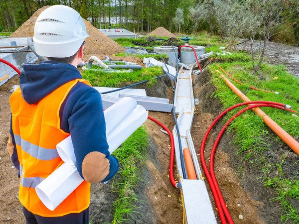 Laying utilities. Man looks at the pipes leading to the well. Builder with drawings next to the water supply. Pipes at the construction site lead. Human in a construction uniform looks at water pipes