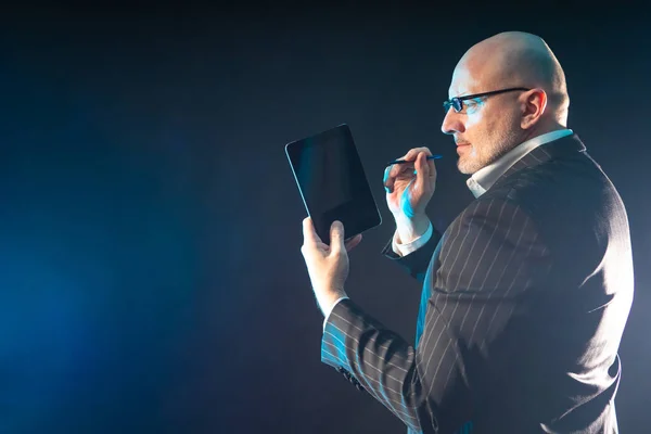 Human with a tablet in his hands. A man in a business suit and glasses. Adult businessman on a dark background. Concept - A man works on a tablet. The gadget is off. Modern businessman