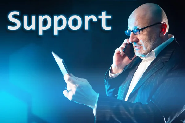 A man in a black suit holds an electronic tablet and calls the support service. Technical support for subscribers. Round-the-clock consulting of clients by phone and online. Hotline.
