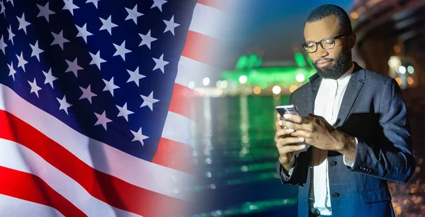 An American reads the news in a mobile phone. An African-American man with a smartphone in his hands on the background of the us flag. An American student looks at the phone screen.