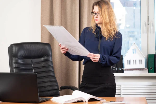 Woman in the office is considering a large file. Girl with a big sheet of paper in her hands. Office manager at the workplace. Work office manager. Girl in the personal account works with documents