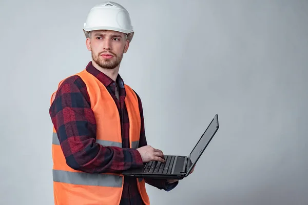 A man in an orange vest and hard hat is holding a laptop. Construction engineer. The architect in the hard hat looks away. Monitoring the progress of construction. Checking all stages of construction.