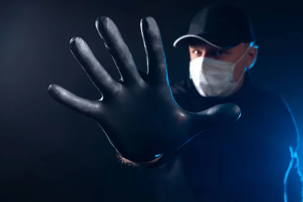 The man with the mask stretched forth his hand in black rubber glove. A hand in a black rubber glove with the fingers spread out in a close-up. Disturbing photo. A black man in a medical mask.