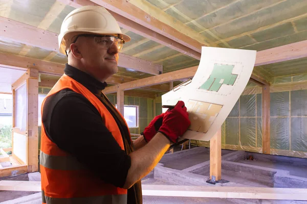 Construction of cottages. A man inside a house under construction. Builder with room plans in hand. Smiling master at the construction site. The man controls the progress of work.