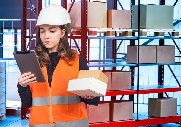 Warehouse. Warehouse logistics. Search for the desired box using an electronic tablet. A girl with boxes in her hands against the background of the warehouse.
