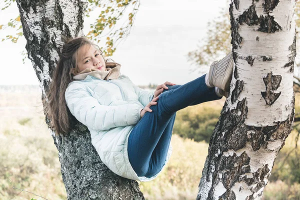 青いダウンジャケットの長い髪を持つ若い女の子は 秋の日に白樺の木に横たわっています — ストック写真