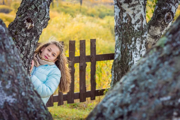 美しいです女の子で長い髪でダウンジャケット秋の日に白樺の列の外を見る. — ストック写真