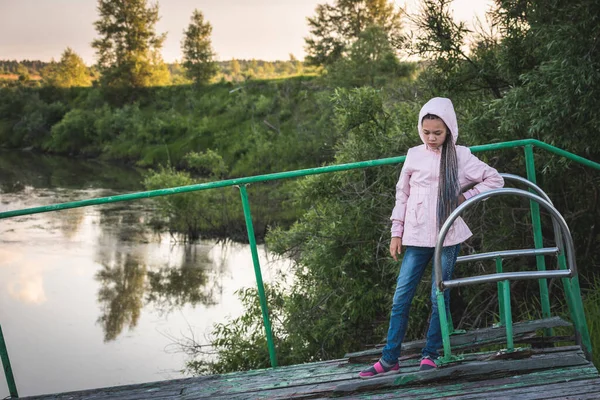 フード付きの夏のジャケットの女の子は桟橋に立っており、日没で悲しいです. — ストック写真