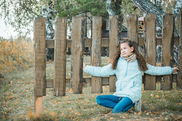Schönes Mädchen in Schal und blauer Daunenjacke hockt an einem kühlen Herbsttag mit Holzzaun im Dorf. — Stockfoto