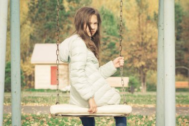 Serin sonbahar gününde oyun alanında salıncak oturan aşağı ceket giymiş Sevimli kız.