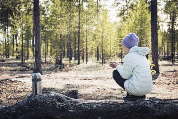 Девушка в светлой куртке и шляпе сидит и отдыхает после прогулки в лесу на змее рядом с термосом чая . — стоковое фото